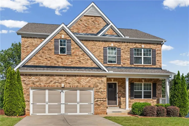 Brick facade with gabled roofline, covered front porch with white columns, attached garage with decorative doors, and established front landscaping