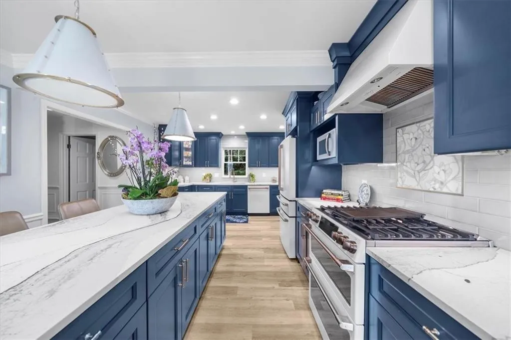 Kitchen featuring blue cabinetry, white appliances, under cabinet range hood, light stone countertops, and tasteful backsplash