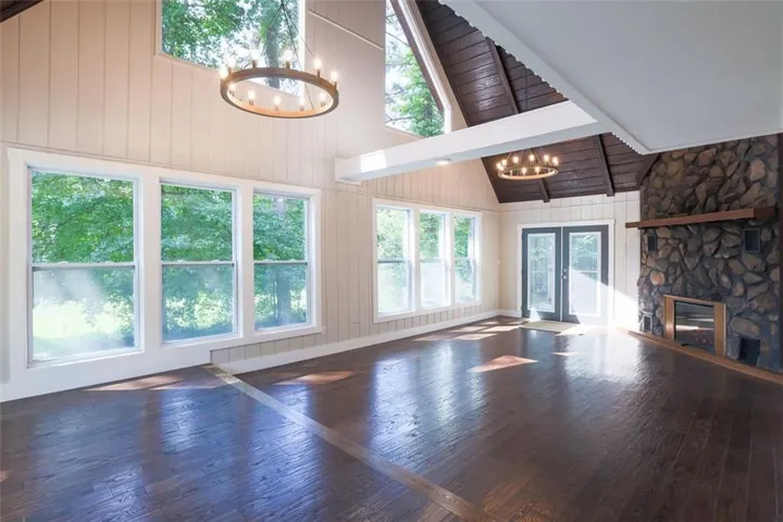 Vaulted ceiling with exposed wood beams, stone fireplace with a wood mantle, wood-finish flooring, multiple large windows, and French doors