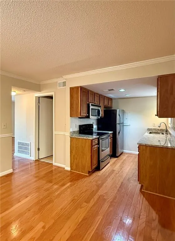 Kitchen with brown cabinets, stainless steel appliances, light wood-style flooring, crown molding, and light stone counters