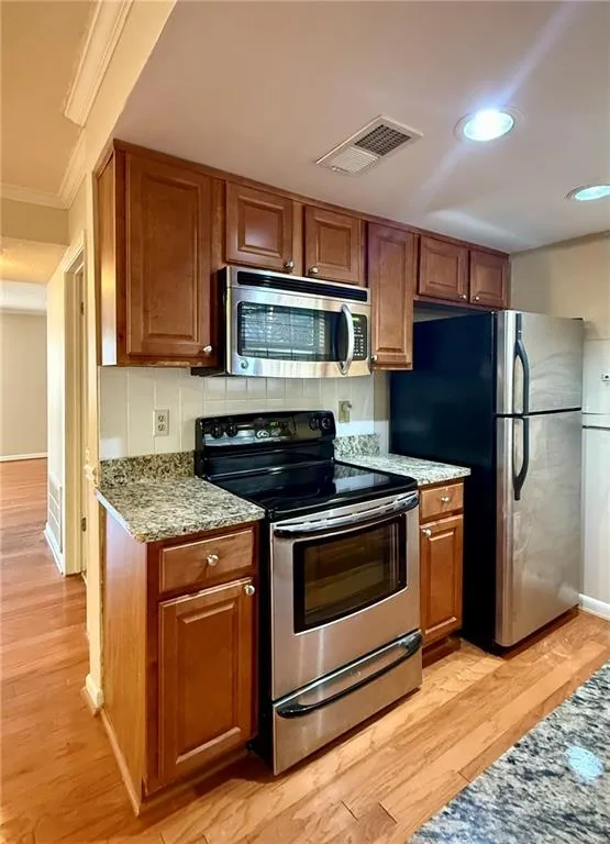 Kitchen featuring stainless steel appliances, brown cabinetry, ornamental molding, light wood finished floors, and light stone counters