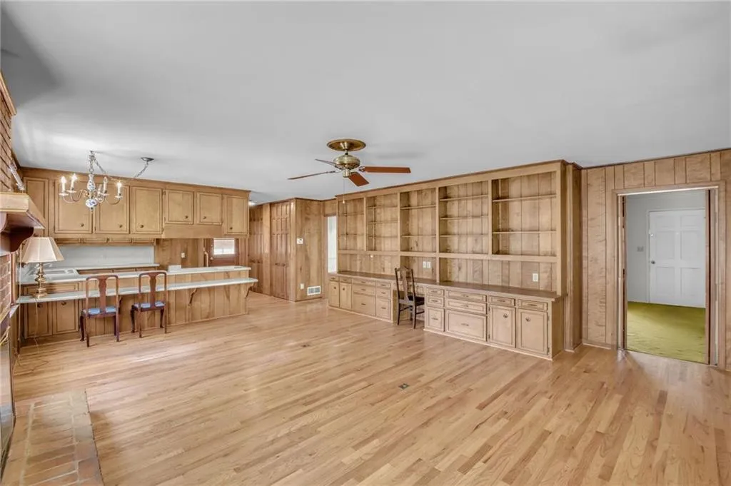 Living room featuring light wood-type flooring, wood walls, built in desk, and ceiling fan with notable chandelier