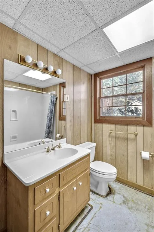 Full bath featuring toilet, wooden walls, vanity, a shower with curtain, and a drop ceiling