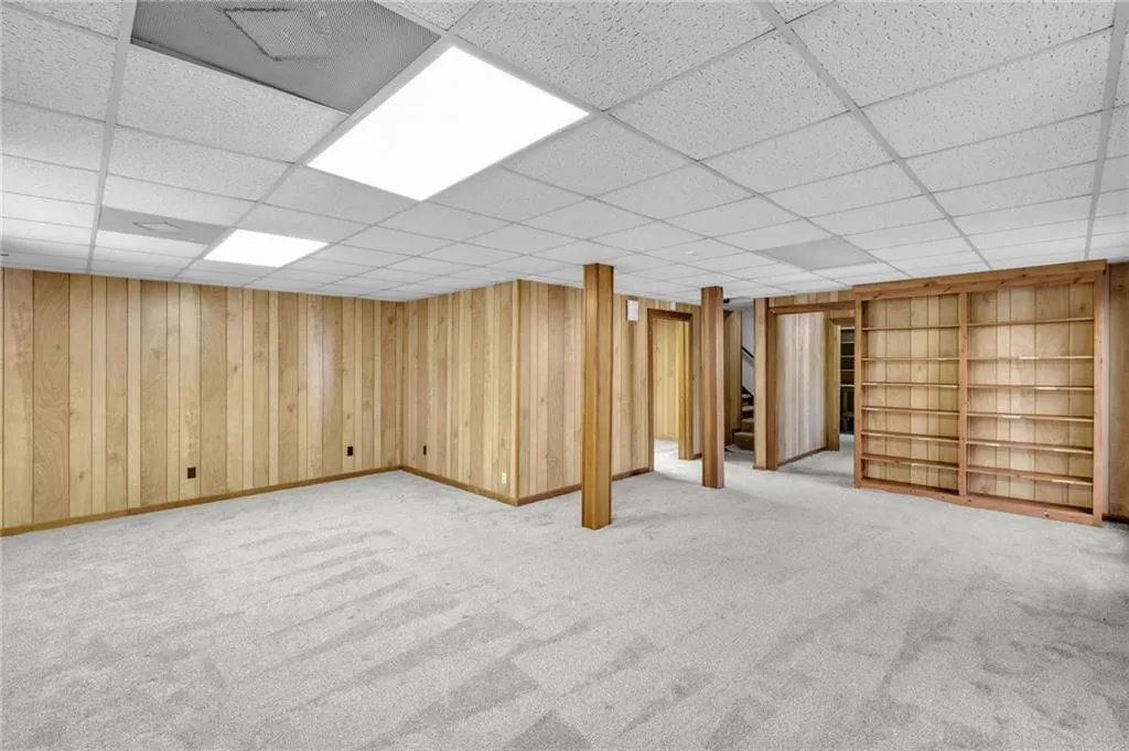Finished below grade area featuring light carpet, wooden walls, baseboards, a drop ceiling, and built in shelves