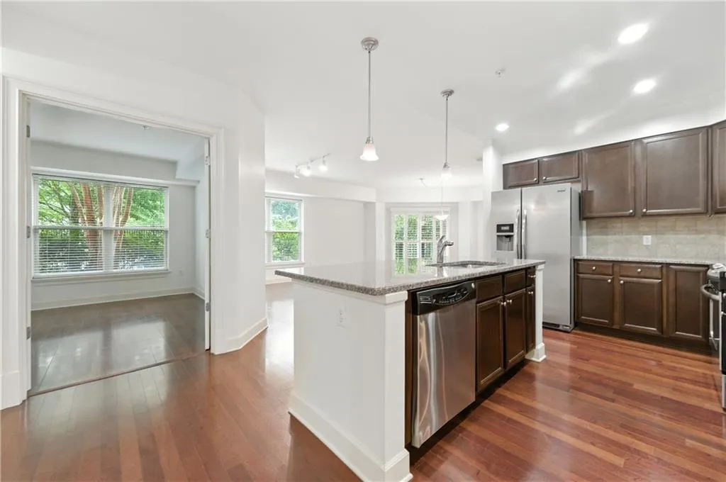 Kitchen featuring backsplash, light stone counters, decorative light fixtures, an island with sink, and dark wood-style floors