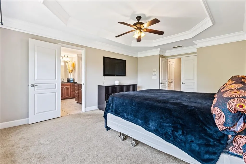 Bedroom with light colored carpet, a raised ceiling, crown molding, ensuite bath, and a ceiling fan