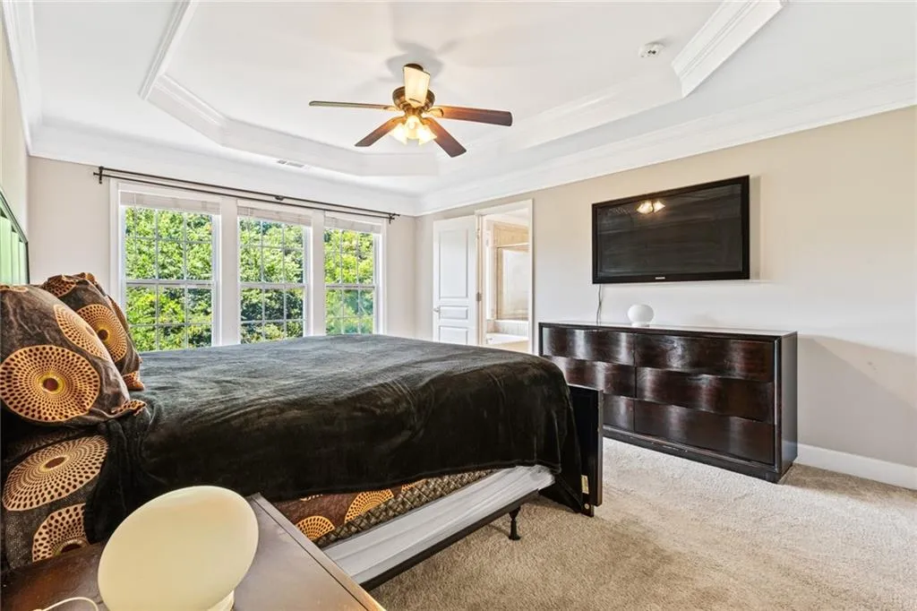 Carpeted bedroom featuring crown molding, a raised ceiling, ensuite bath, and a ceiling fan