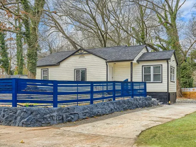 Yellow siding exterior with dark trim, black shingle roof, and a blue slatted privacy fence