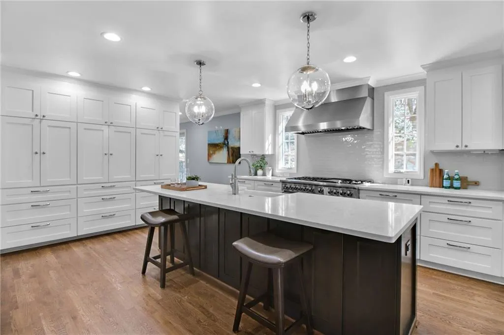Kitchen featuring wall chimney range hood, sink, white cabinetry, hanging light fixtures, and a center island with sink