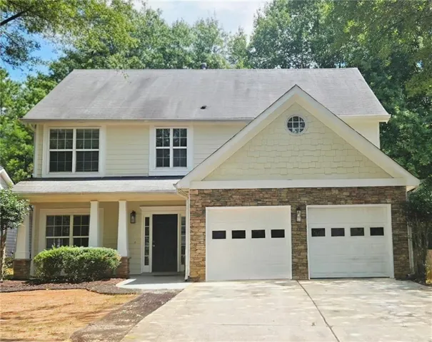 Two-story residence featuring a stone-clad facade, dual garage doors with window inserts, a covered front porch with pillar supports, and a prominent gable with decorative shingle siding and a circular window