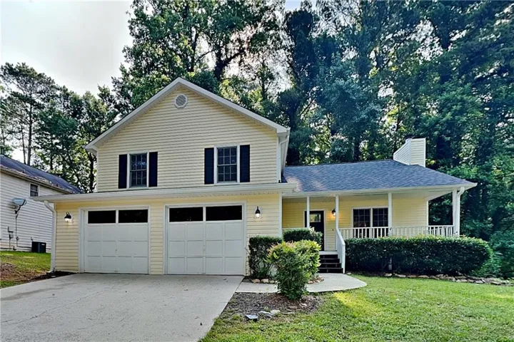 Light yellow siding exterior with dark shutters, white garage doors, and a covered front porch