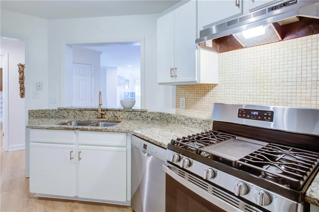 Kitchen with appliances with stainless steel finishes, under cabinet range hood, white cabinetry, backsplash, and light stone counters
