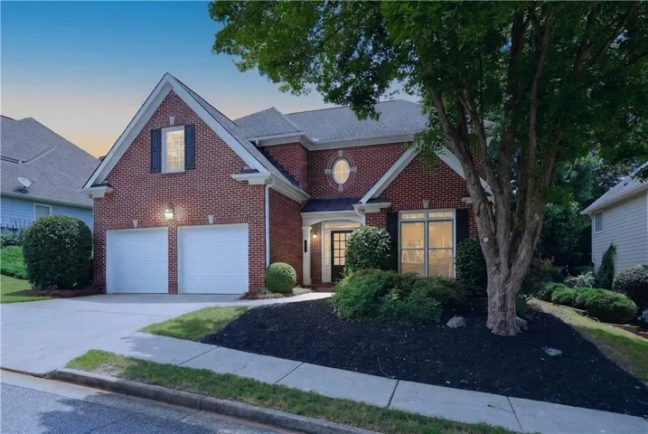 Brick exterior with a gabled roofline, featuring a two-car garage, a main entry with a covered porch, and mature trees enhancing the landscaping
