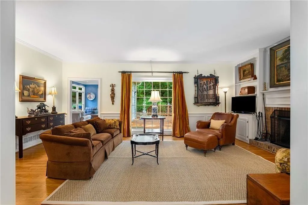 Family room/Den featuring wood finished floors, a brick fireplace, ornamental molding, a wainscoted wall, and a large window with view of backyard and deck.