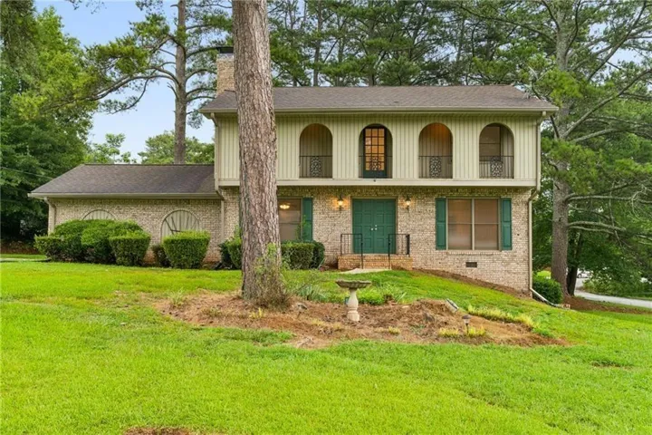 Brick and siding exterior featuring arched window details, prominent green double entry doors, green window shutters, and a sloped front lawn with mature trees