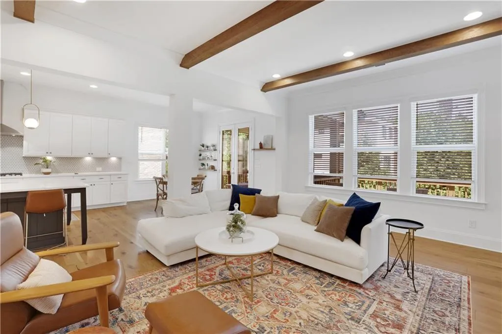 Open-concept living space featuring wood-finish flooring, exposed ceiling beams, recessed lighting, white cabinetry, and multiple windows with blinds