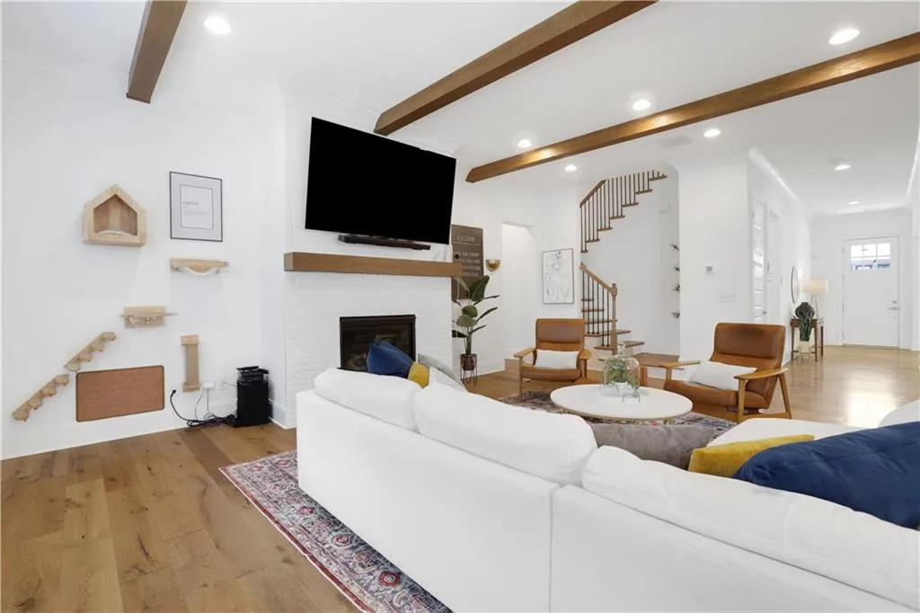 Spacious living area featuring exposed wood beams, wood-finish flooring, a white brick fireplace with a wood mantel, a visible staircase with wood treads and banister, and recessed lighting
