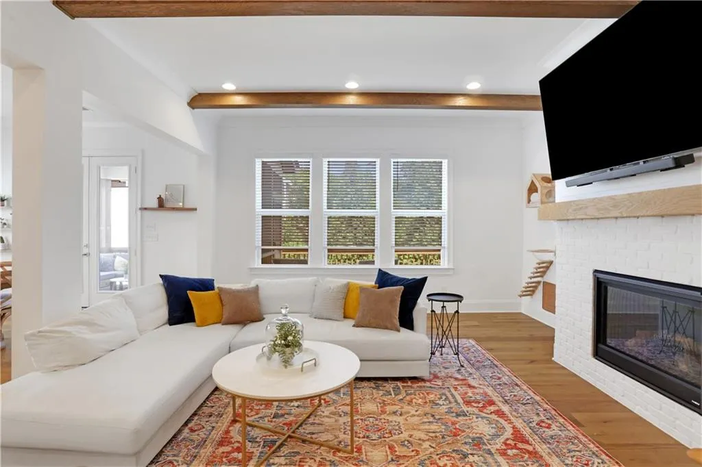 Spacious living area featuring exposed wood ceiling beams, a white brick fireplace with a wood-finish mantel, recessed lighting, and wood-finish flooring
