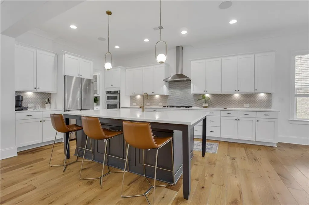 Spacious kitchen featuring an expansive center island with seating, a stainless steel range hood, double wall ovens, a tiled backsplash, and wood-finish flooring