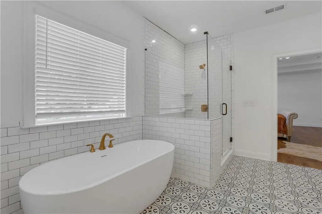 Bathroom with a freestanding soaking tub featuring brass fixtures, a glass-enclosed shower with subway tile surround, patterned floor tiling, a window with blinds, and recessed lighting