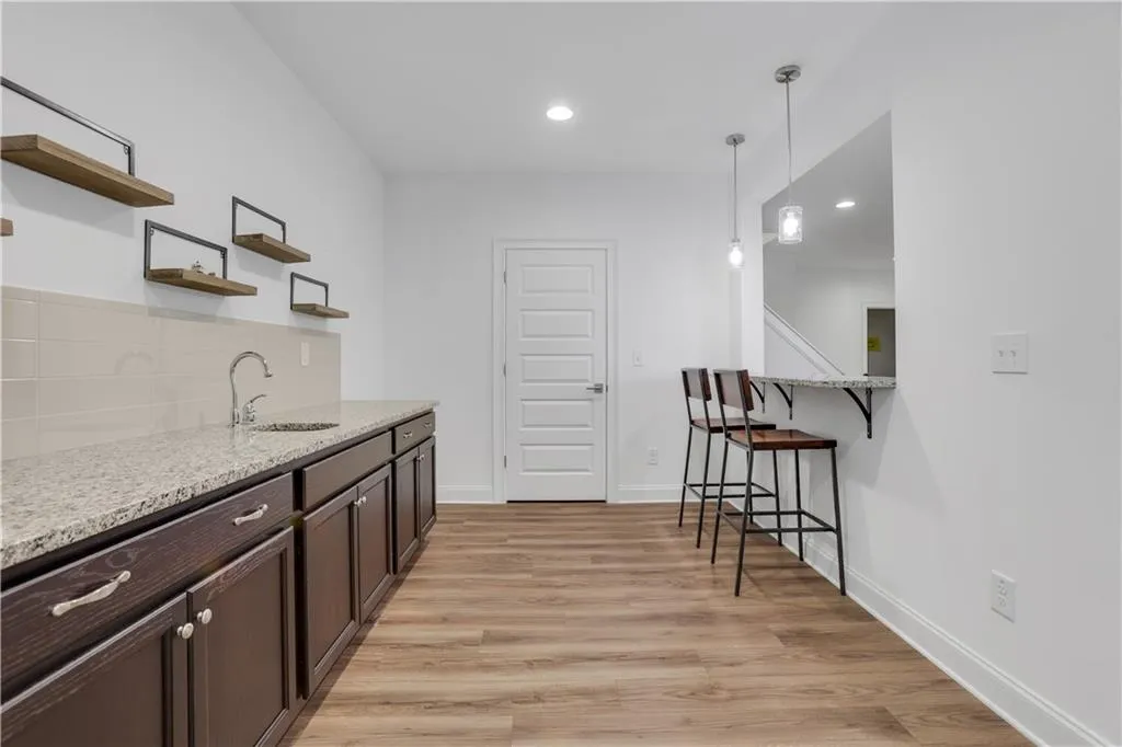 Wet bar featuring wood-finish flooring, dark wood cabinetry with chrome hardware, granite-style countertops, and a subway tile backsplash