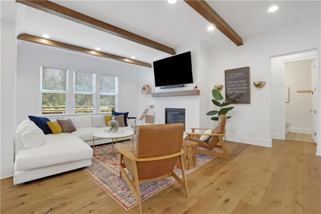 Spacious living area featuring wood-finish flooring, exposed ceiling beams, recessed lighting, and a white brick fireplace with a wood mantel