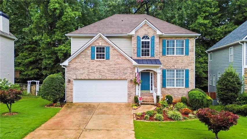 Brick Colonial two-story home featuring a two-car garage, blue shutters and trim accents, arched entryway with brick detailing, manicured front lawn with mature landscaping including boxwoods and flowering shrubs, and a concrete driveway