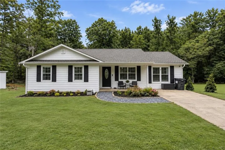 White siding exterior with black shutters, dark shingle roof, paved driveway, and established landscaping