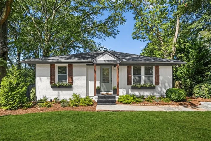 White brick exterior with dark wood shutters, front porch with wood columns, dark gray roof, and landscaped garden beds