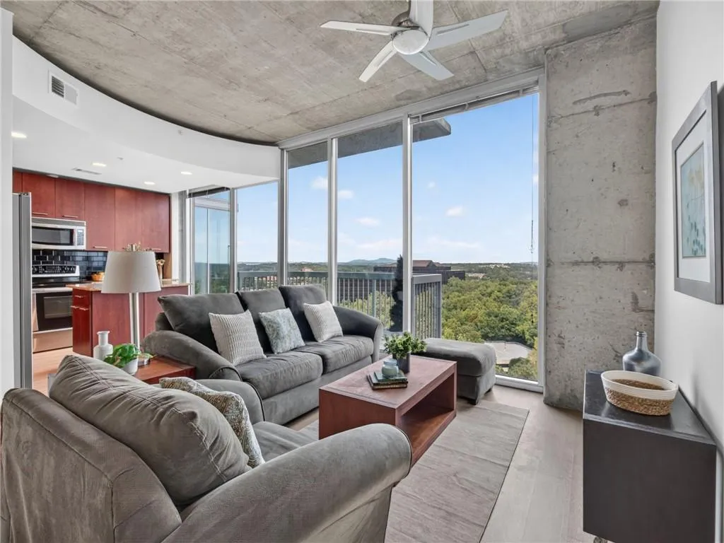 Expansive window wall with balcony access, wood-finish flooring, exposed concrete ceiling, and a kitchen featuring stainless steel appliances and dark wood cabinetry