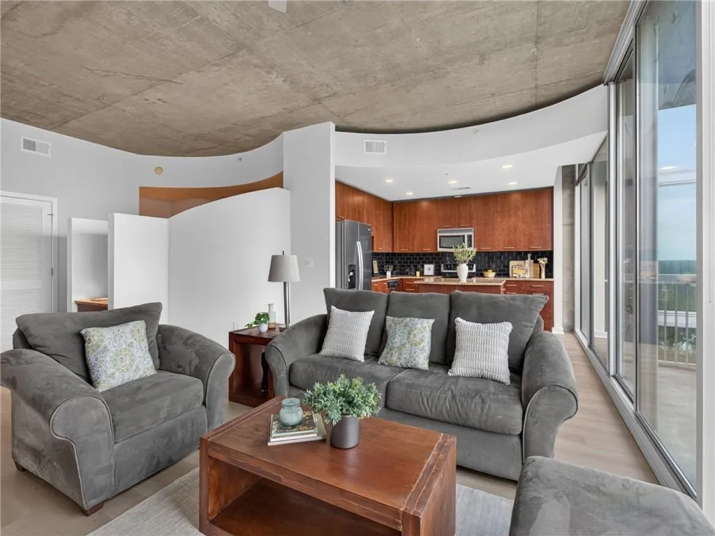 Expansive floor-to-ceiling windows, exposed concrete ceiling, contemporary curved wall, wood-finish flooring, and built-in kitchen cabinetry. Living room with a view to the kitchen.