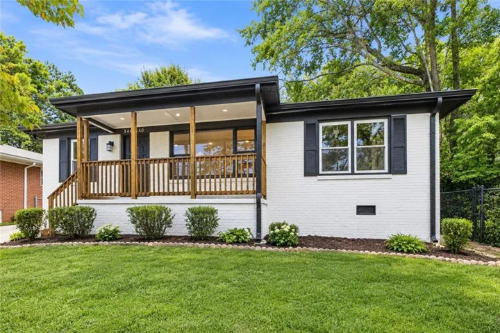 White brick exterior with black trim, a covered front porch featuring wood columns and railing, front lawn, and mature trees