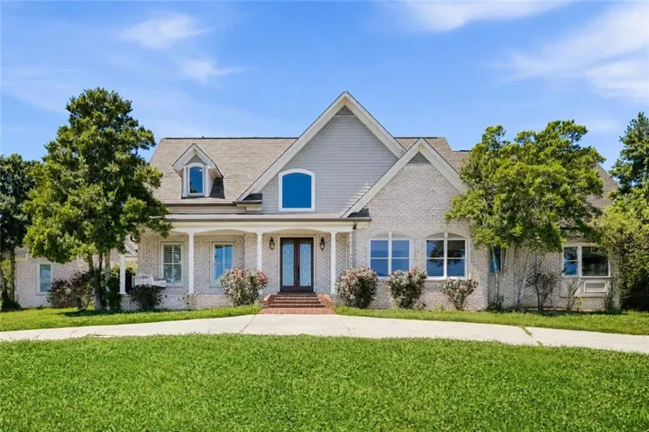 White brick exterior with a welcoming covered porch, double entry doors, dormer window, and mature landscaping