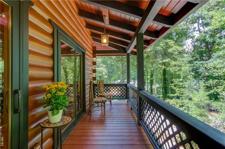 Covered deck with wood-finish flooring and exposed beam ceiling, featuring lattice railing and large windows framing the surrounding forest landscape