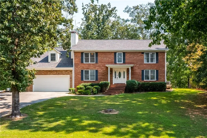 Traditional brick exterior featuring a white columned entryway, dark shutters, an attached two-car garage, dormer window, and mature landscaping