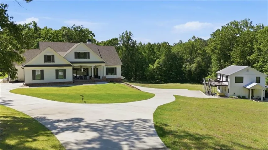 Expansive estate featuring a main residence with a covered front porch, a separate auxiliary building with an exterior deck, a paved circular driveway, and mature trees providing a natural backdrop