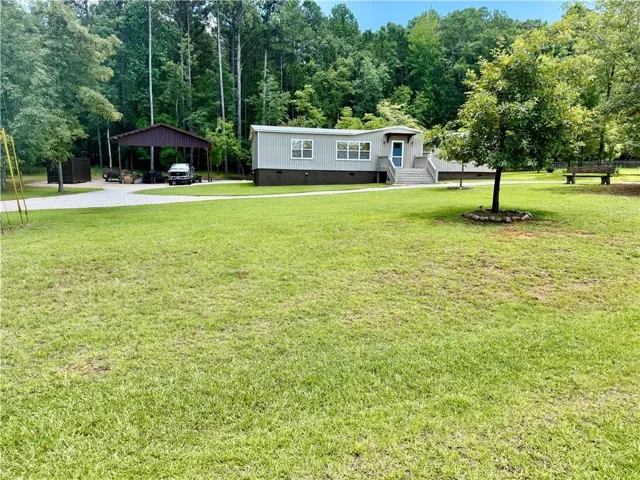 Expansive grassy yard area with a gravel driveway, a detached carport, mature trees, and a gray-sided residence featuring a front entry deck with stairs