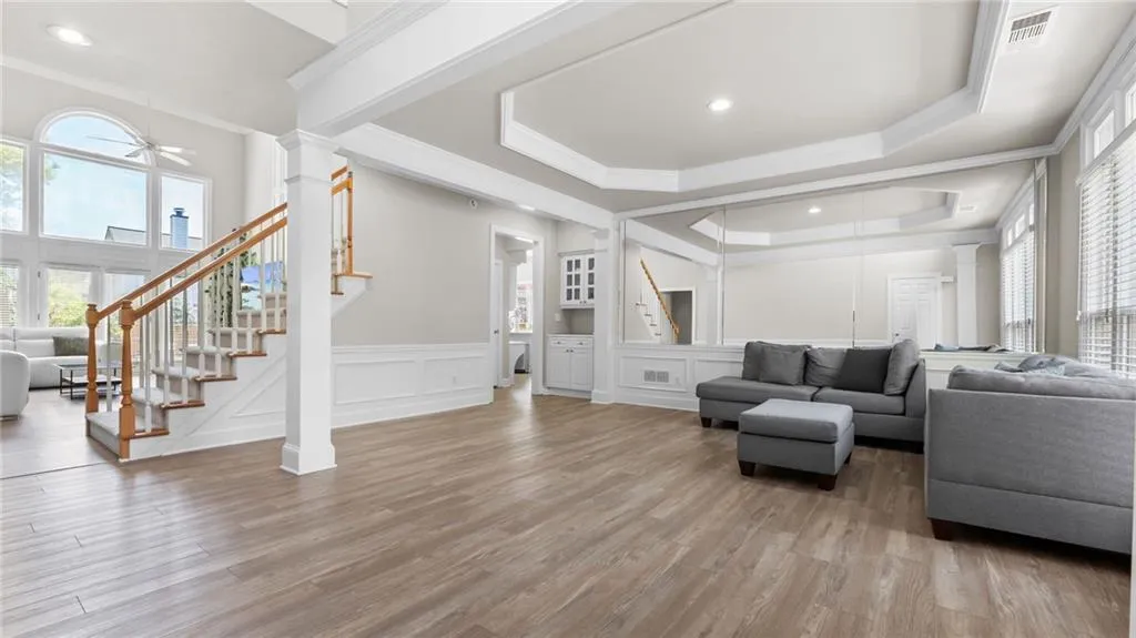 Living area featuring a ceiling fan, stairway, ornate columns, a raised ceiling, and a decorative wall