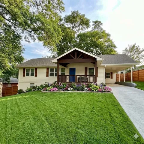Virtual Image, 
Cream-colored siding exterior with dark wood shutters, covered front porch with wood railings, attached carport, concrete driveway, and mature landscaping with a flower bed