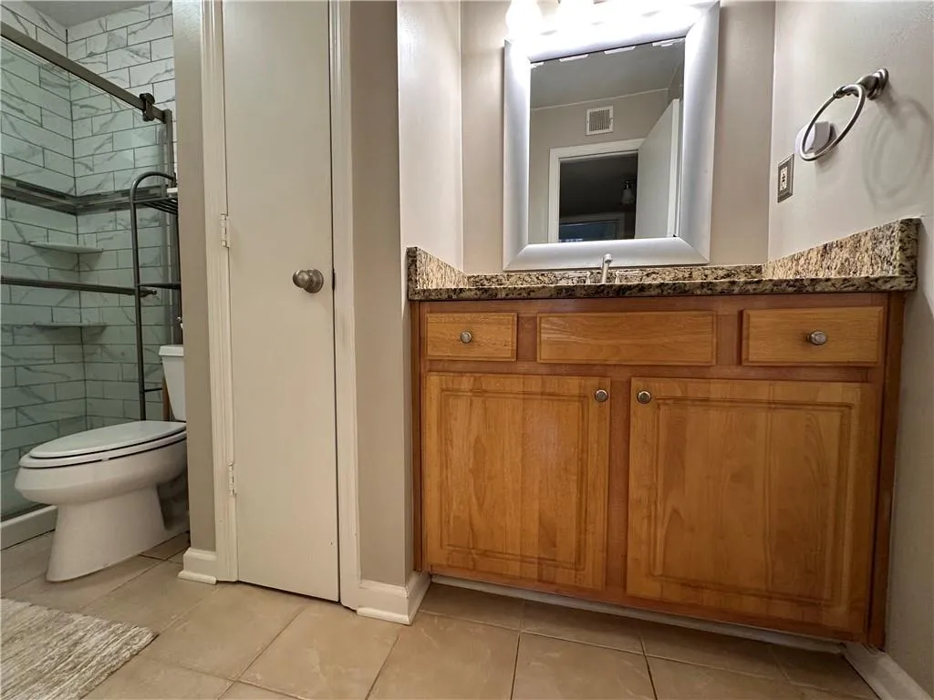 Bathroom featuring a wood-finish vanity with a granite-style countertop, a framed mirror with integrated lighting, a toilet, and a shower enclosure with subway tile surround and built-in shelving