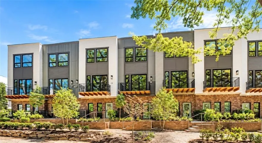 Modern multi-story townhome featuring a stone and panel exterior, large windows, individual balconies with iron railings, and a landscaped front terrace