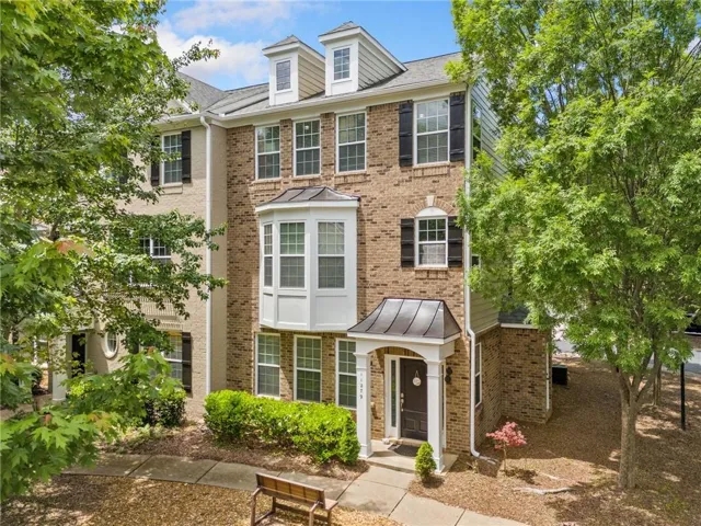 Brick facade townhome with bay window, prominent entry porch, dormer windows, and established landscaping