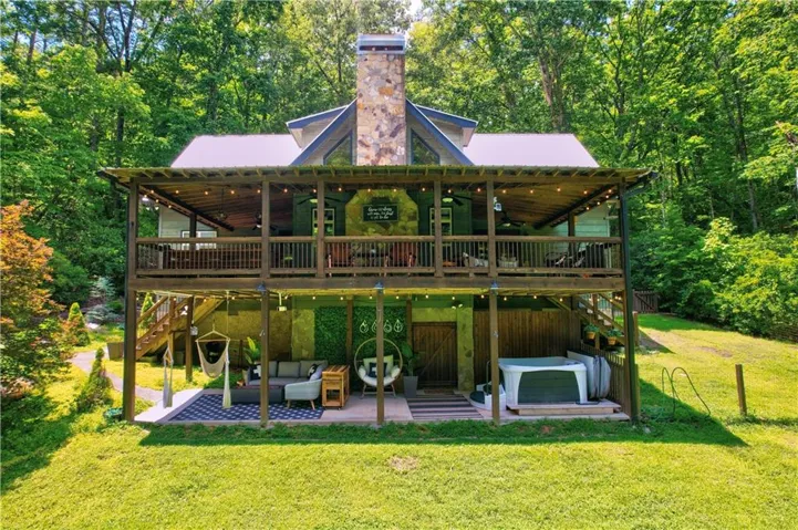 Two-story structure featuring a stone chimney, metal roof, upper-level covered balcony, lower-level covered patio, and attached hot tub