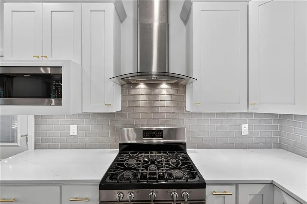 Kitchen with stainless steel appliances, wall chimney exhaust hood, white cabinets, backsplash, and light stone countertops