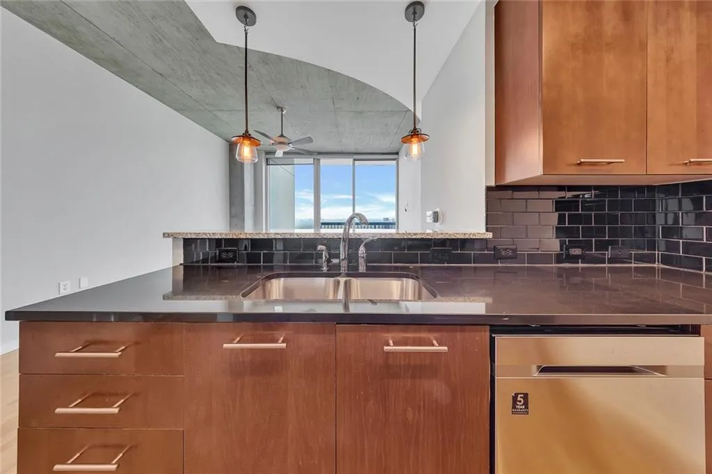 Kitchen featuring dark countertops, a stainless steel sink with a gooseneck faucet, black subway tile backsplash, and wood-finish cabinetry with linear hardware