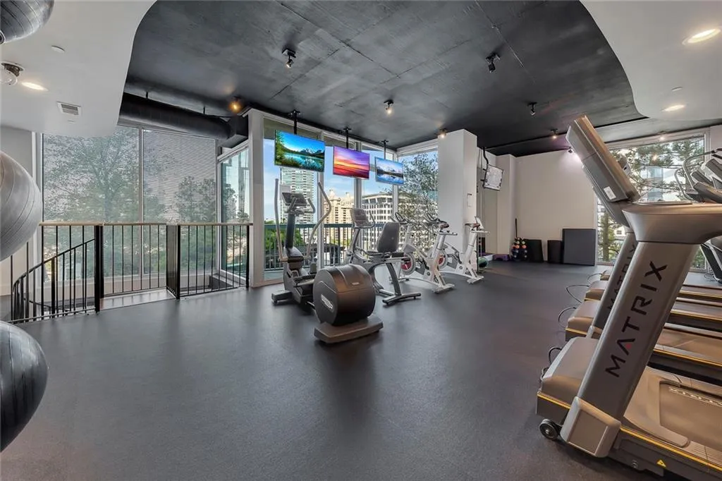 Fitness area featuring rubber flooring, exposed ceiling with track lighting, multiple screens, and large windows