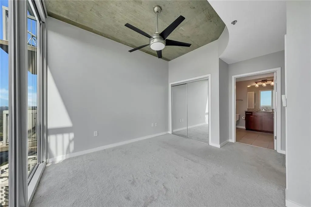 Floor-to-ceiling windows, gray carpeting, exposed concrete ceiling, ceiling fan, and mirrored closet doors