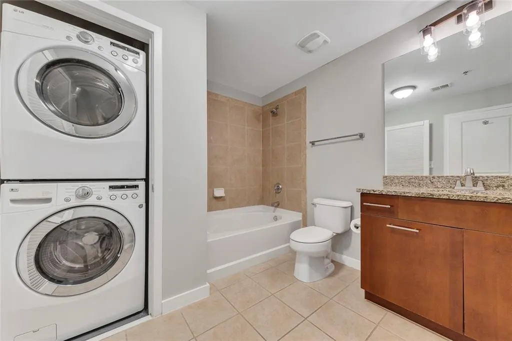 Stacked washer and dryer, tile flooring, vanity with stone-finish countertop, full bathtub with tile surround, and an overhead light fixture