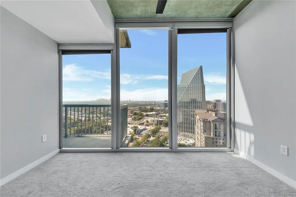Full-height windows with city views, balcony access, concrete ceiling, and light-toned carpeting