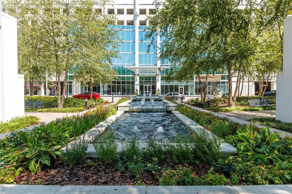 Modern building exterior with glass facade, multiple fountains, mature trees, and landscaped planters